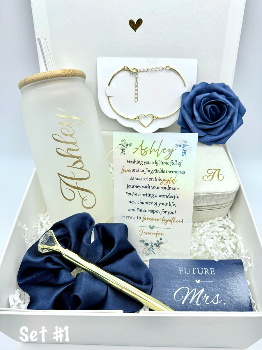 navy blue themed gift box for a bride. set 1 includes: glass tumbler personalized with the name of the bride, metal heart bracelet, foam rose, personalized make up box, metal pen, scrunchie, named "wishing you a lifetime full of life..." card. a "future mrs." card. the box has white paper filler.