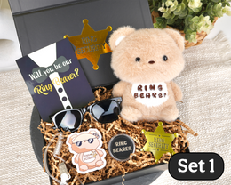 A styled gift set in an open black box filled with shredded brown paper, featuring a beige teddy bear plush with a 'RING BEARer!' patch, a black card reading 'Will you be our Ring Beaver?', black sunglasses, a beige bear-shaped pin, a round black 'RING BEARER' coin, and gold sheriff star pins. The box sits on a beige fabric surface with decorative props in the background.