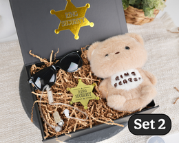 A ring security gift set in a black box with shredded paper filler, featuring a plush teddy bear with a 'Ring  BEARER' sign, black sunglasses, a gold sheriff star badge, and small jewelry pieces. A 'Set 2' label is visible in the bottom right corner.