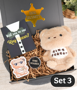 A ring bearer gift set in a black box filled with brown paper shred, featuring a plush tan bear with a white patch reading 'RING BEARER,' a black card that says 'Will you be our Ring Bearer?', a gold sheriff star pin, a small round pin, and a die-cut sticker. A black 'Set 3' label appears in the lower right corner.