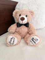 ring bearer beige colored plush bear. one foot personalization is "ring bearer" with hearts and the other foot has rings, the girls name and date of the wedding. all in black colored iron-on vinyl. the bear has a matching bowtie on its neck, matching the color of the text