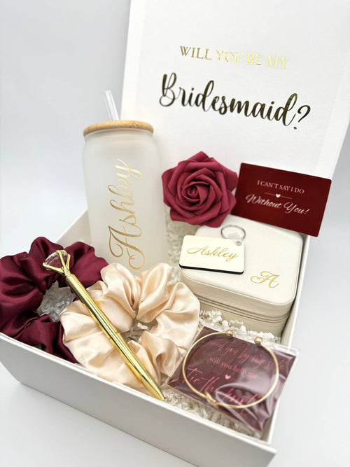 Burgundy bridesmaid proposal gift box with “Will you be my bridesmaid?" on the inner lid, magnetic lid. with personalized glass cup with a straw, jewelry box, a burgundy scrunchie and a cream colored scrunchie, a gold colored metal diamond pen, a faux rose head, a "I can't say I do without you" card, a personalized keychain, a metal bracelet, a "I need you by my side, will you help me tie the knot" card.