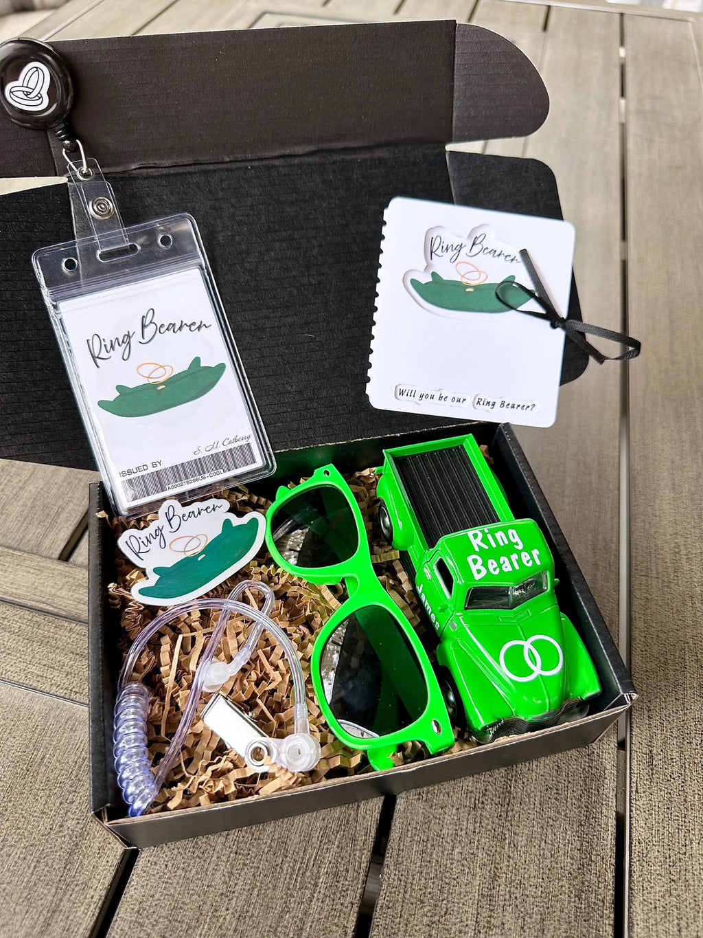 open black box that is a ring bearer proposal gift box. items included are a green ring bearer ID badge, green "will you be our ring bearer" proposal card tied with a bow. green sunglasses, green die-cast car personalized with "ring bearer" on the roof, rings on the hood, date of the wedding on the back and name of the ring bearer on the doors. also included are an agent ear piece and a green "ring bearer" sticker. box comes with brown paper filler