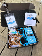 open black box that is a ring bearer proposal gift box. items included are a light blue ring bearer ID badge, light blue "will you be our ring bearer" proposal card tied with a bow. light blue sunglasses, light blue die-cast car personalized with "ring bearer" on the roof, rings on the hood, date of the wedding on the back and name of the ring bearer on the doors. also included are an agent ear piece and a light blue "ring bearer" sticker. box comes with brown paper filler