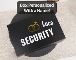 A black rectangular personalized gift box with the words 'Luca SECURITY' and a gold interlocking rings graphic, placed on a beige textured fabric surface. A black banner at the top reads 'Box Personalized With a Name!'.