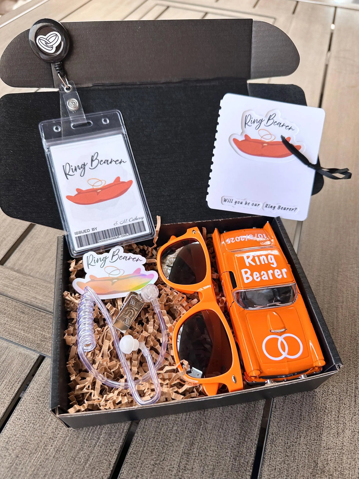 open black box that is a ring bearer proposal gift box. items included are an orange ring bearer ID badge, orange "will you be our ring bearer" proposal card tied with a bow. orange sunglasses, orange die-cast car personalized with "ring bearer" on the roof, rings on the hood, date of the wedding on the back and name of the ring bearer on the doors. also included are an agent ear piece and an orange "ring bearer" sticker. box comes with brown paper filler