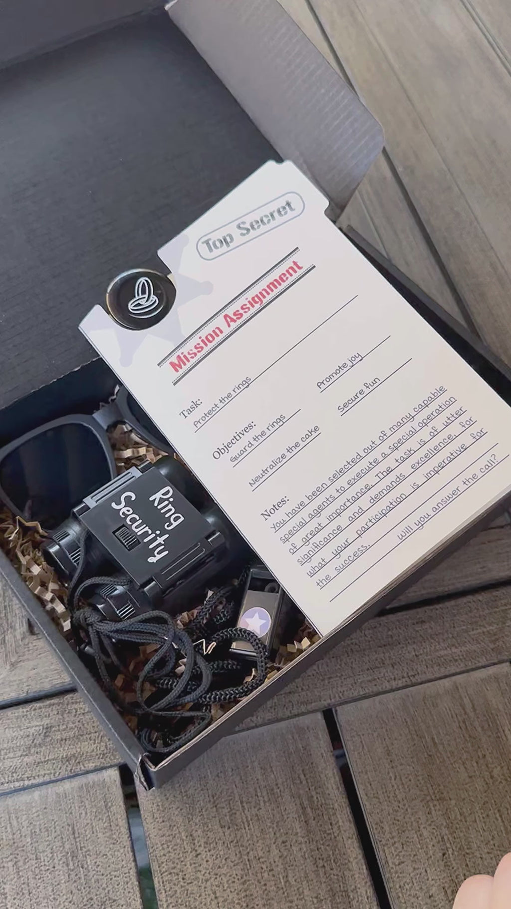 detailed video showcasing each product for the full set of ring security proposal box. open box, set 1 includes a ring security "mission assignment" card with a ring security agent id badge, sunglasses, play binoculars, agent earpiece, black whistle, "ring security" sticker, personalized black car with a name. the inside of the box is filled with Kraft colored paper filler.