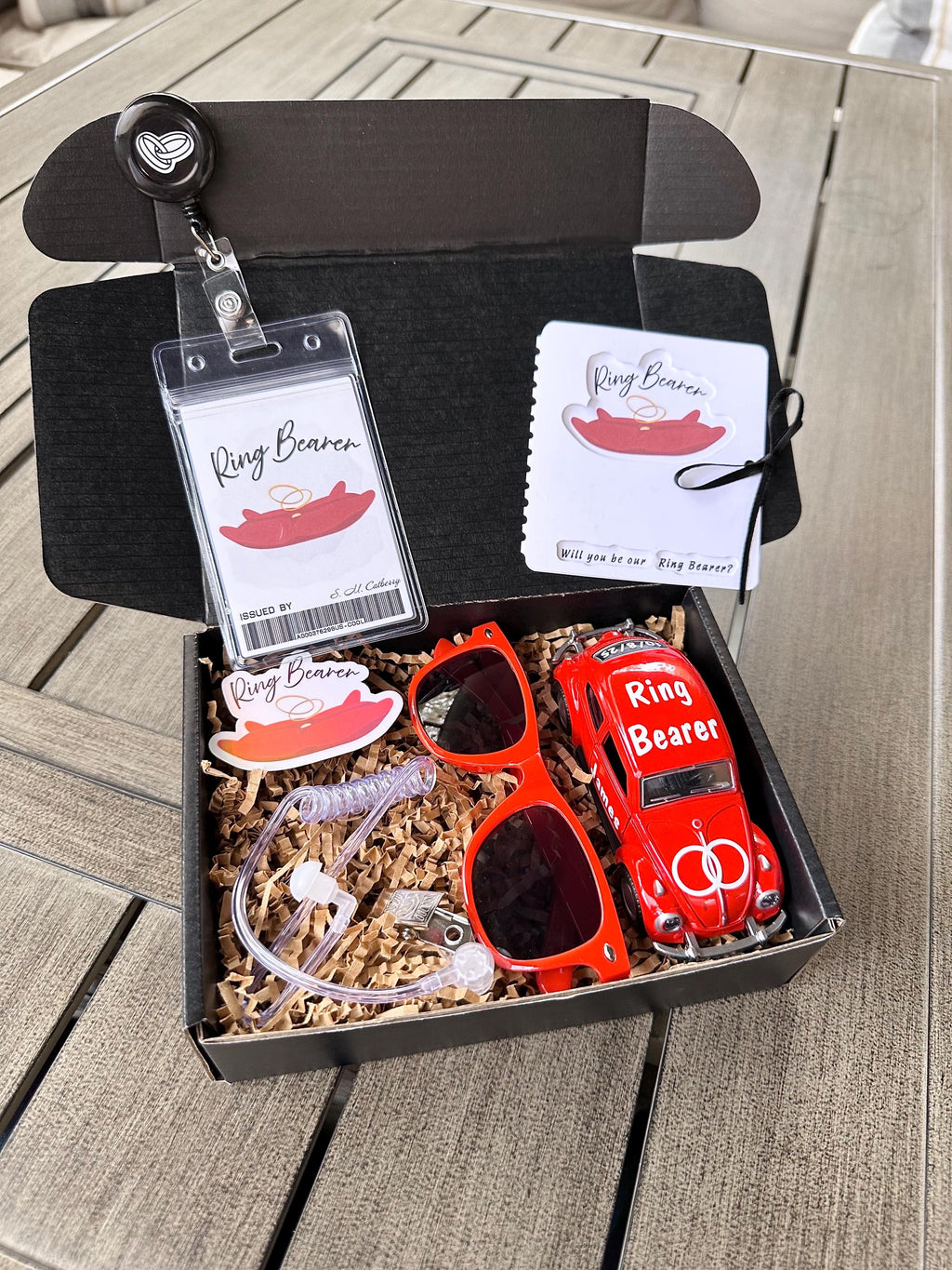 open black box that is a ring bearer proposal gift box. items included are a red ring bearer ID badge, red "will you be our ring bearer" proposal card tied with a bow. red sunglasses, red die-cast car personalized with "ring bearer" on the roof, rings on the hood, date of the wedding on the back and name of the ring bearer on the doors. also included are an agent ear piece and a red "ring bearer" sticker. box comes with brown paper filler