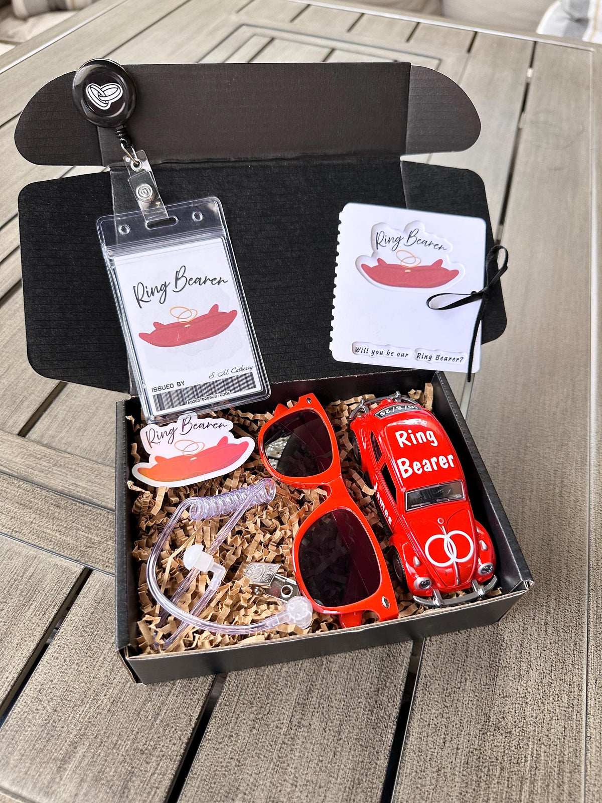 open black box that is a ring bearer proposal gift box. items included are a red ring bearer ID badge, red "will you be our ring bearer" proposal card tied with a bow. red sunglasses, red die-cast car personalized with "ring bearer" on the roof, rings on the hood, date of the wedding on the back and name of the ring bearer on the doors. also included are an agent ear piece and a red "ring bearer" sticker. box comes with brown paper filler