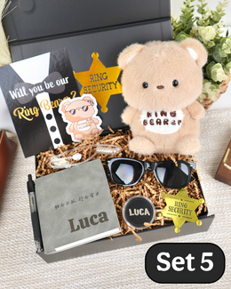 A curated gift set box labeled 'Set 5' containing a plush teddy bear with a 'Ring Bearer' patch black sunglasses, a custom name card reading 'Luca', a black card that says 'Will you be our Ring Bear?', a gold sheriff star badge, and a black box lid, arranged in brown paper shred.