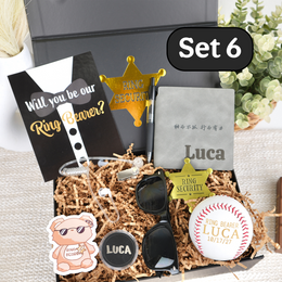 A personalized ring bearer gift set arranged in a box filled with shredded paper. The set includes a black card reading 'Will you be our Ring Bearer?', a gold badge-shaped pin, a gray notebook with the name Luca, sunglasses, a white baseball with red stitching and 'RING BEARER LUCA' text, a small bear sticker, and other matching accessories.
