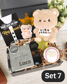 A gift set arranged in a black box with a magnetic lid with shredded paper filler, featuring a plush teddy bear with 'Ring Bearer' text stitched on it, a agent earpiece, black sunglasses, a white baseball with 'Luca' and a ring design, a black card, and gold star-shaped accessories. A black "will you be our ring bearer" card